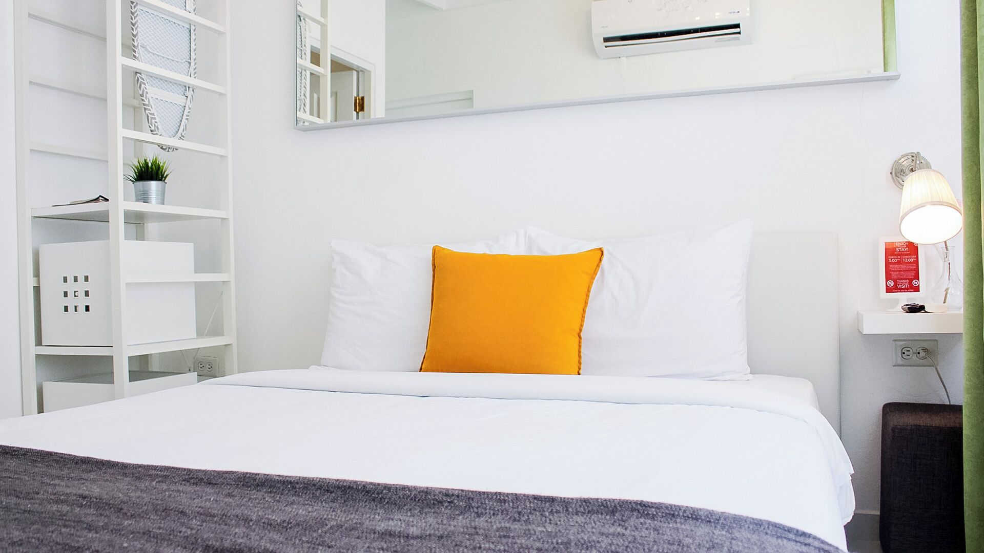 Quarto de hotel com cama, paredes brancas, prateleira com caixas organizadoras, uma pequena planta e ar condicionado fixado na parede.