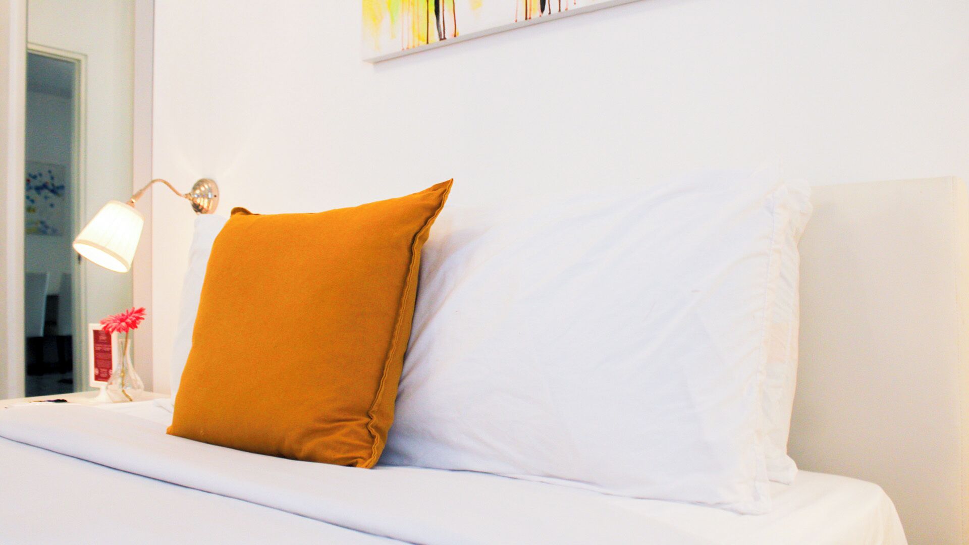 Een bed met witte en mosterdkleurige kussens, een bedlampje en een vaas met bloemen.