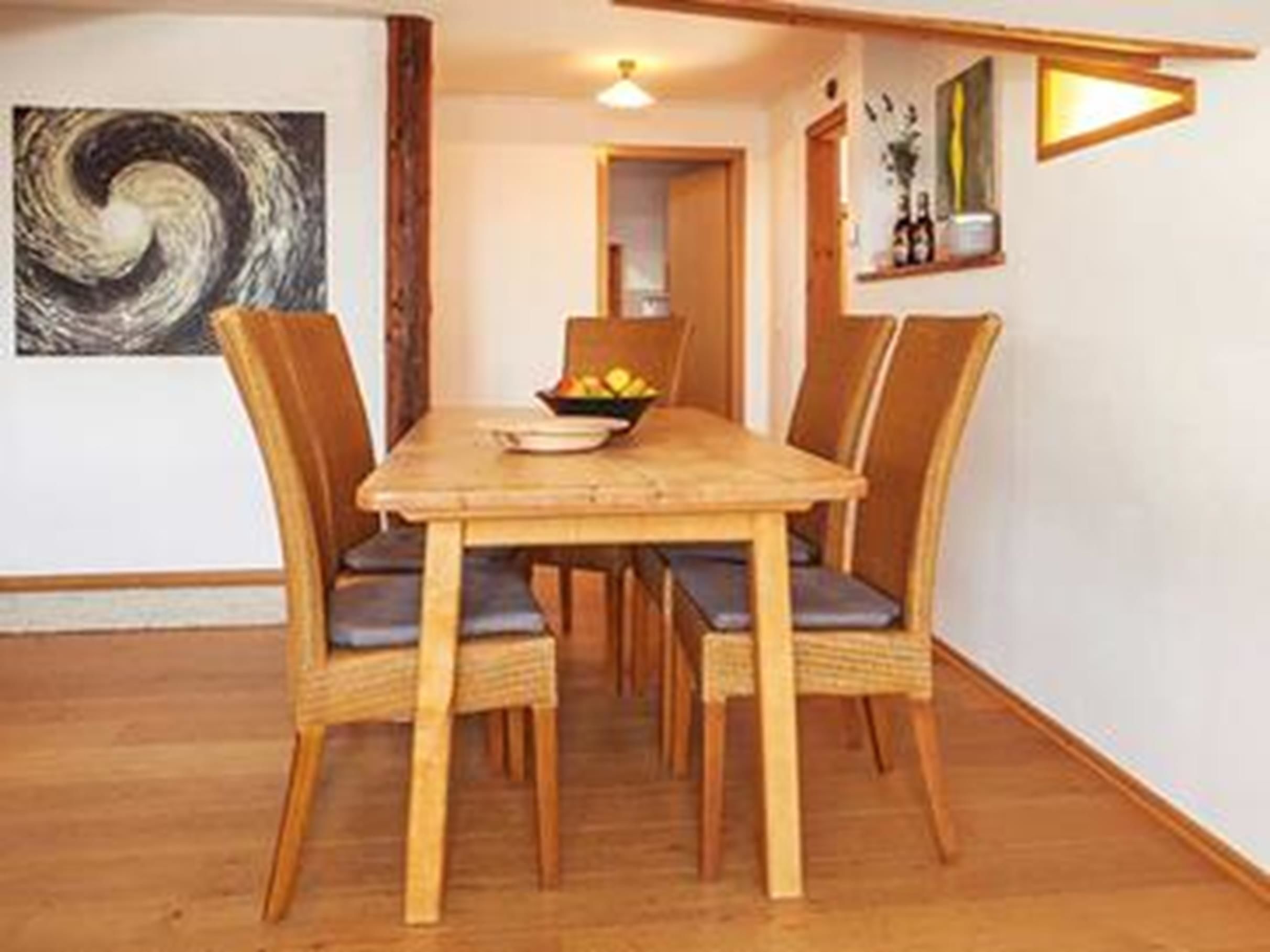 Uma área de refeições com mesa de madeira e quatro cadeiras, uma tigela de frutas e um quadro na parede.