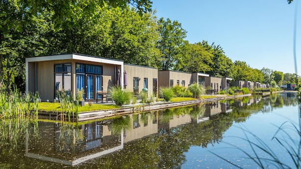 A row of modern, single-story buildings with large windows, situated beside a calm water body, reflecting the structures and surrounding greenery. - EuroParcs Bad Hoophuizen (Hulshorst)