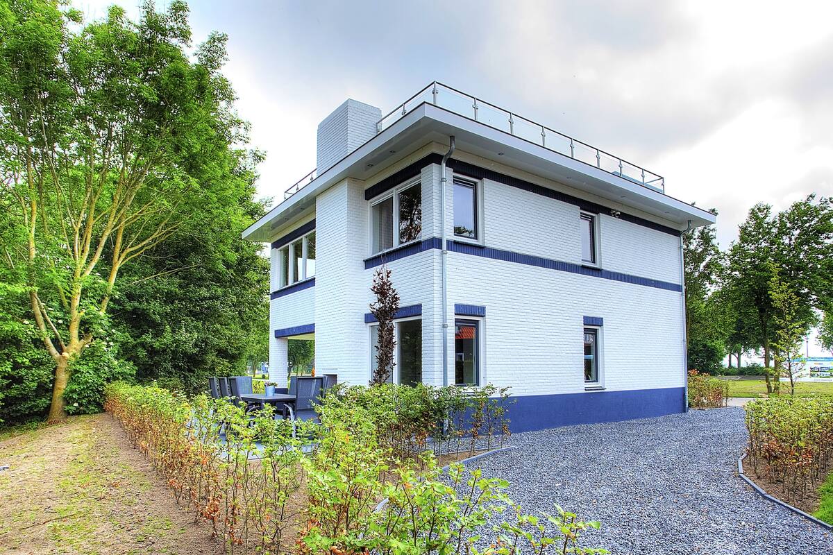 Un edificio moderno de dos plantas con fachada blanca y azul, rodeado de abundante vegetación y un sendero de grava.