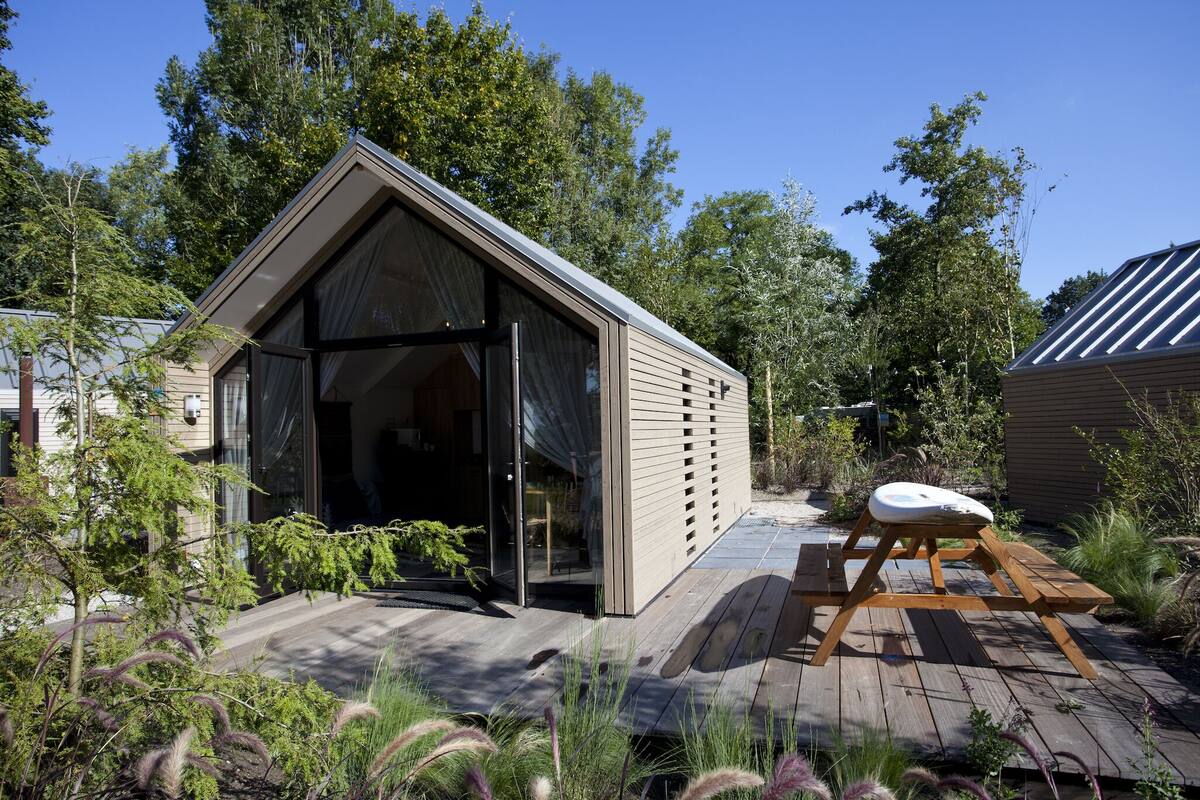 Una cabaña moderna con techo inclinado, puerta de cristal y terraza de madera con banco.