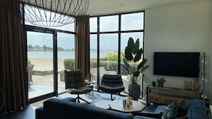 A modern living room with a large window overlooking a body of water, a comfortable sofa, a chair, and a flat-screen TV. - EuroParcs Bad Hoophuizen (Hulshorst)