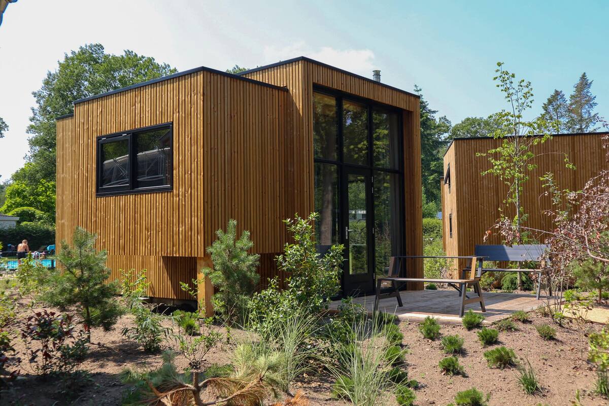 Una cabaña moderna de madera con amplios ventanales, rodeada por un jardín con diversas plantas y una terraza de madera.