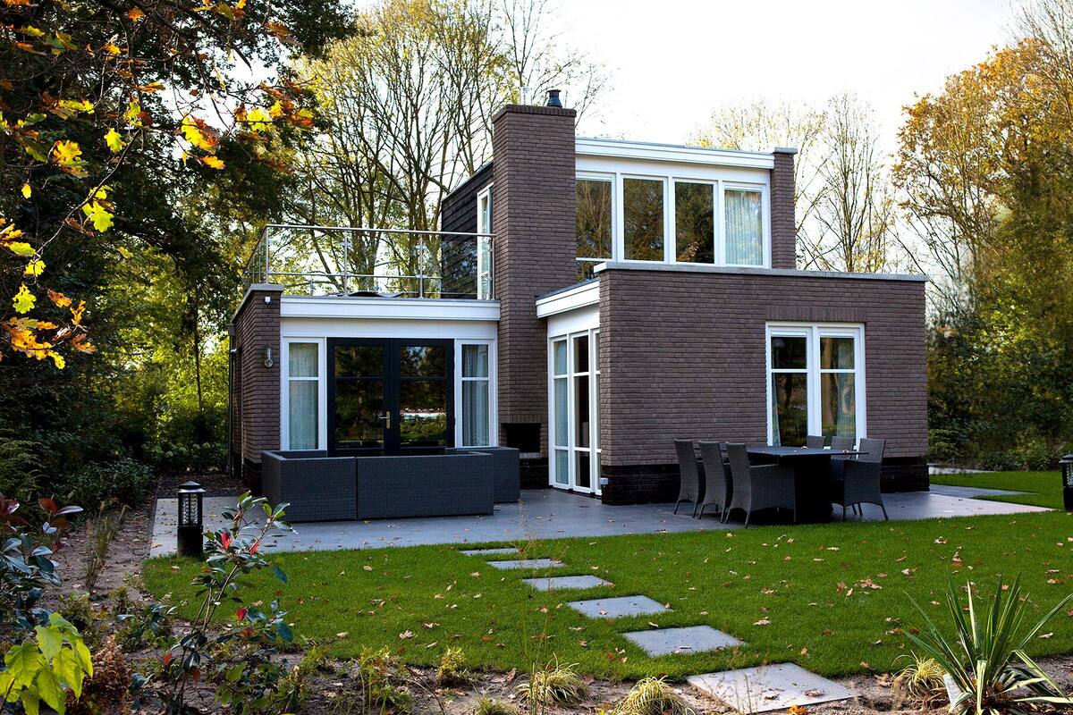 Una casa moderna de dos pisos con fachada de ladrillo, ventanas amplias y un jardín cuidado con patio y mobiliario exterior.