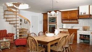A dining area with a wooden table and chairs, a kitchen with wooden cabinets, and a spiral staircase. - Ferienwohnungen Schencking (Reit im Winkl)