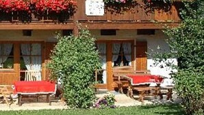A traditional chalet with a wooden facade, two balconies with red flowers, and a small garden area with a bench and table. - Ferienwohnungen Schencking (Reit im Winkl)