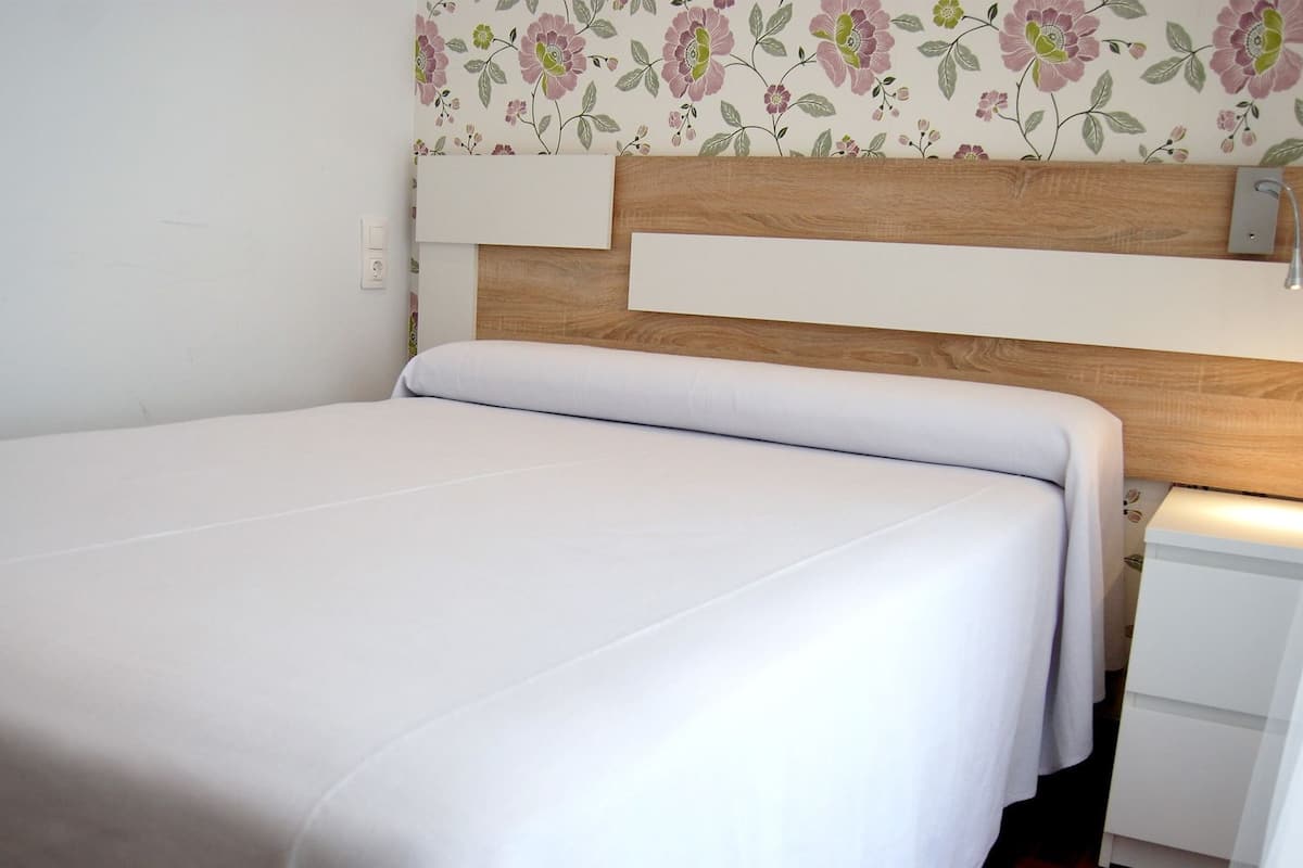 Una cama individual con cabecera de madera, una mesita de noche blanca y una pared con papel tapiz floral.