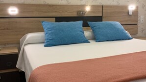 Un letto rifatto con cura, con una coperta bianco-pesco, due cuscini blu e una testiera in legno con illuminazione integrata.