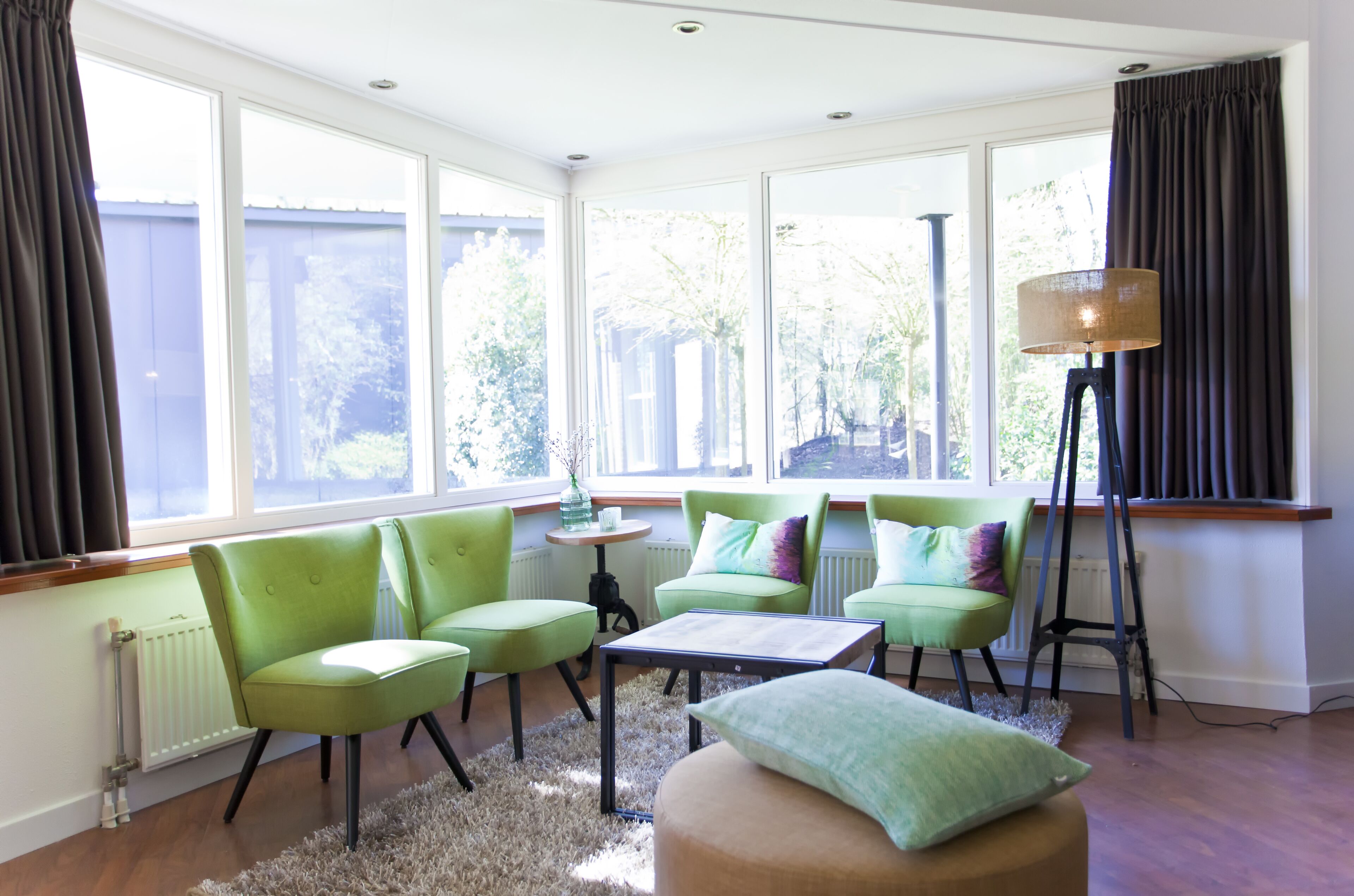 Una habitación luminosa con ventanas amplias, sillas verdes, una mesita y una lámpara de pie.