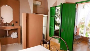 Une chambre avec un lit, une salle de bain avec un lavabo et une porte verte donnant sur un espace extérieur aménagé avec des chaises.