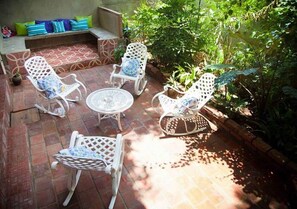 A patio with four white wicker chairs and a small round table, surrounded by a brick floor and lush greenery. - Casa Gardenia's (Trinidad)