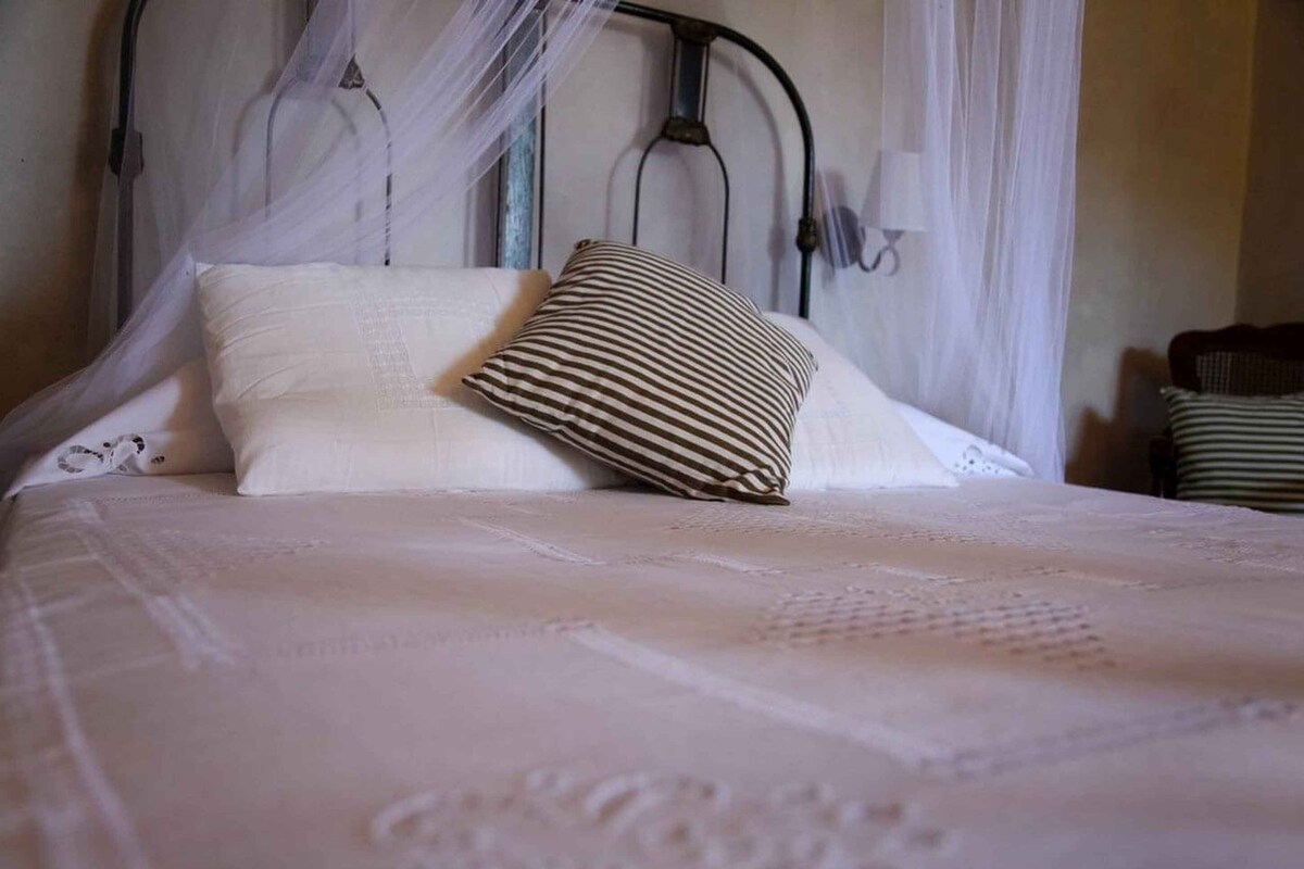 Un lit bien fait, avec un couvre-lit blanc et un oreiller à rayures, une tête de lit en métal et un rideau blanc.