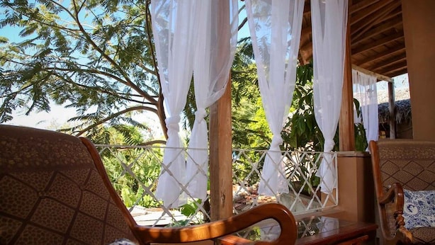 Une chaise en bois avec un coussin à motifs, une petite table et une vue sur un arbre à travers une fenêtre à treillis ornée de rideaux blancs.