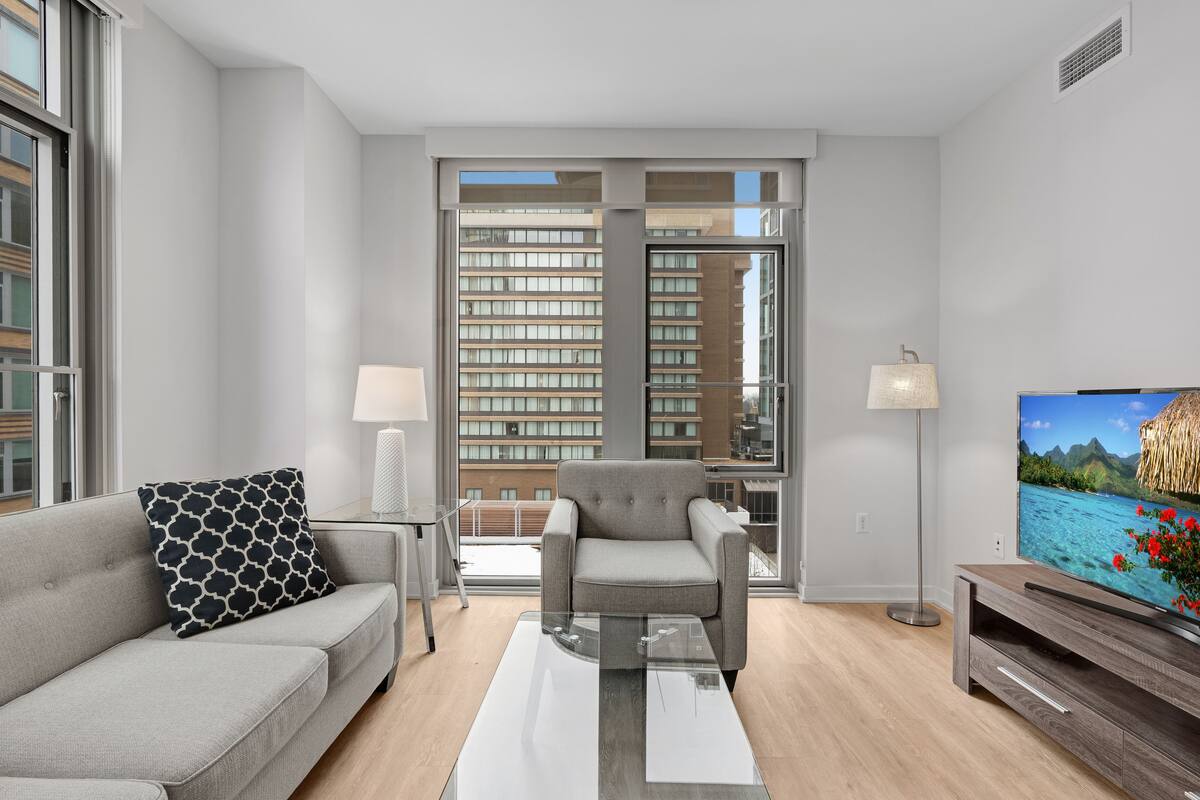 A modern living room with a grey sofa, a patterned throw pillow, a coffee table, a TV on a wooden stand, and a large window with a city view.