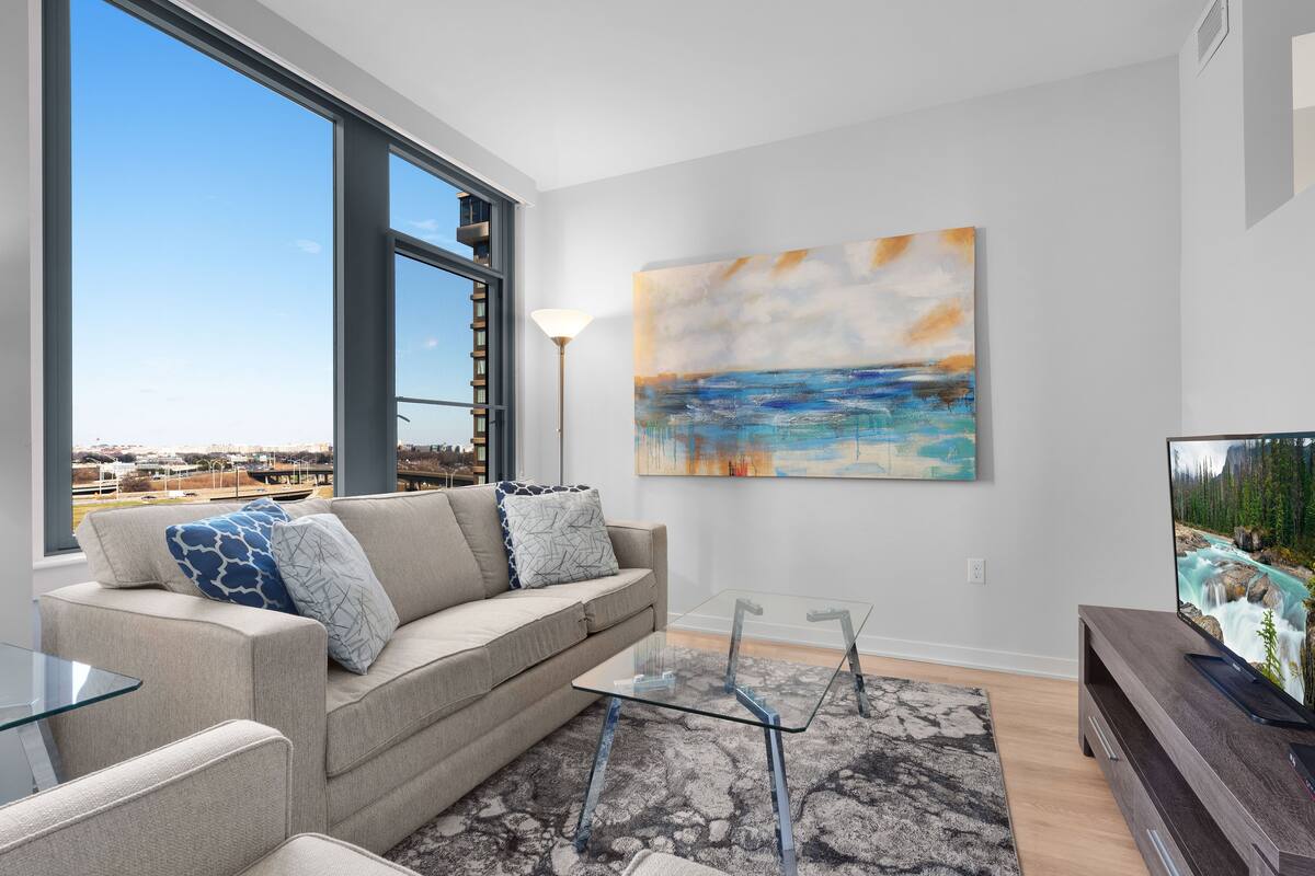 A modern living room with a large window, a sofa with patterned cushions, a glass coffee table, a painting on the wall, and a TV on a wooden stand.