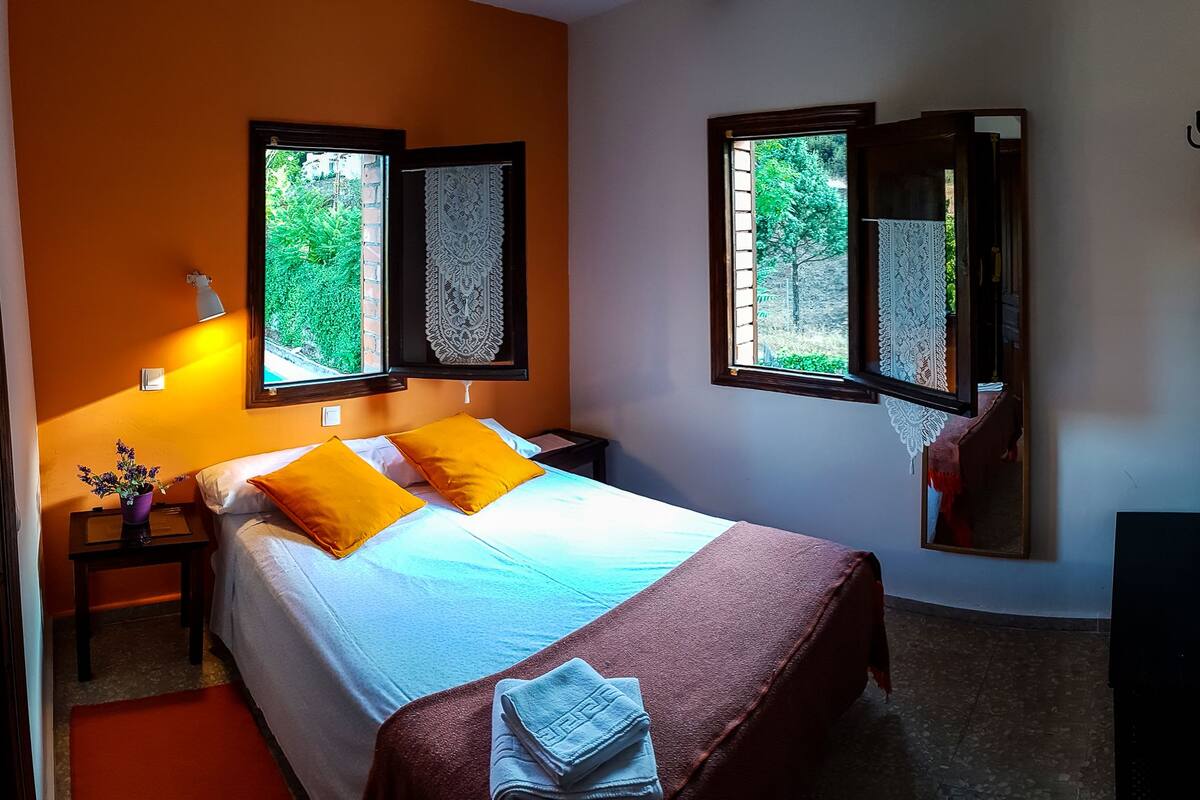 A bedroom with a bed, two windows, a nightstand, a lamp, and a mirror.
