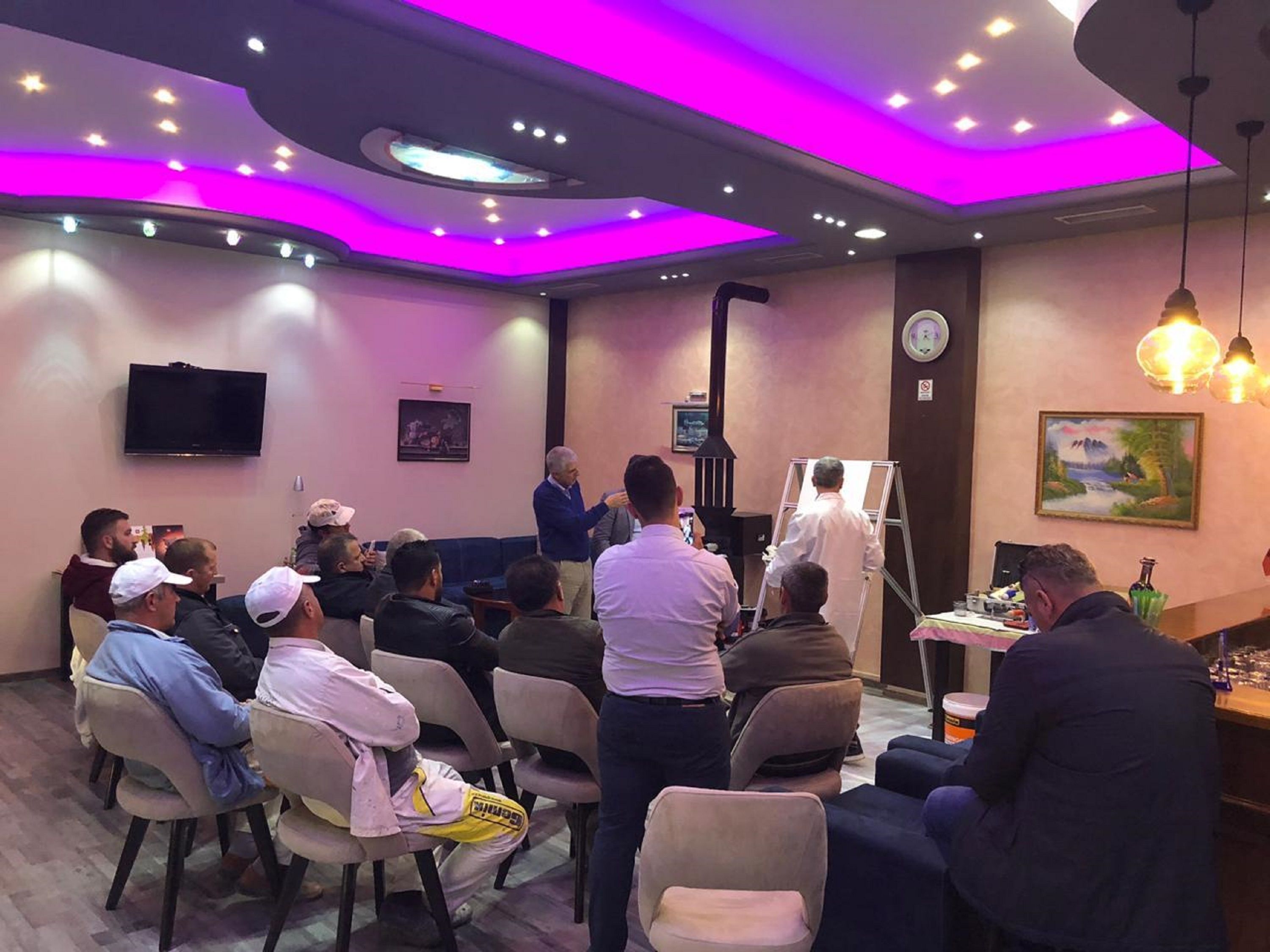 Ein Konferenzraum mit sitzenden und stehenden Personen, einem an der Wand befestigten Fernseher und einer an die Wand gelehnten Leiter.