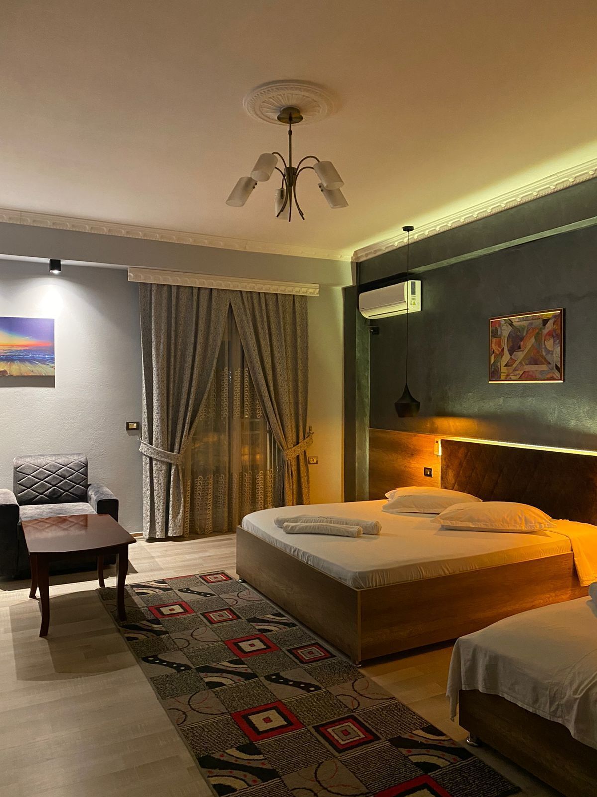 Ein Hotelzimmer mit Bett, Sessel, Tisch, einem gemusterten Teppich, Deckenleuchte und Klimaanlage.