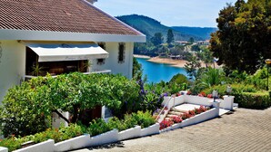 Un edificio con tejado de baldosas, un jardín con diversas plantas y flores, y una vista a una playa y montañas al fondo.