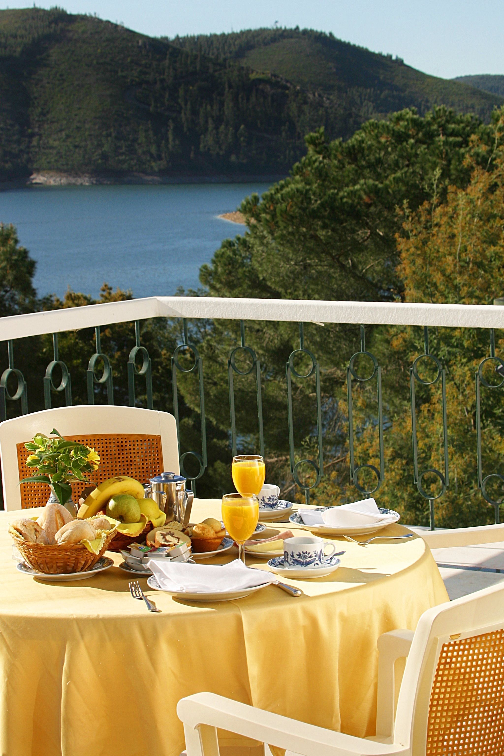 A table set with breakfast items, including bread, fruit, and juice, overlooking a lake and forested hills.