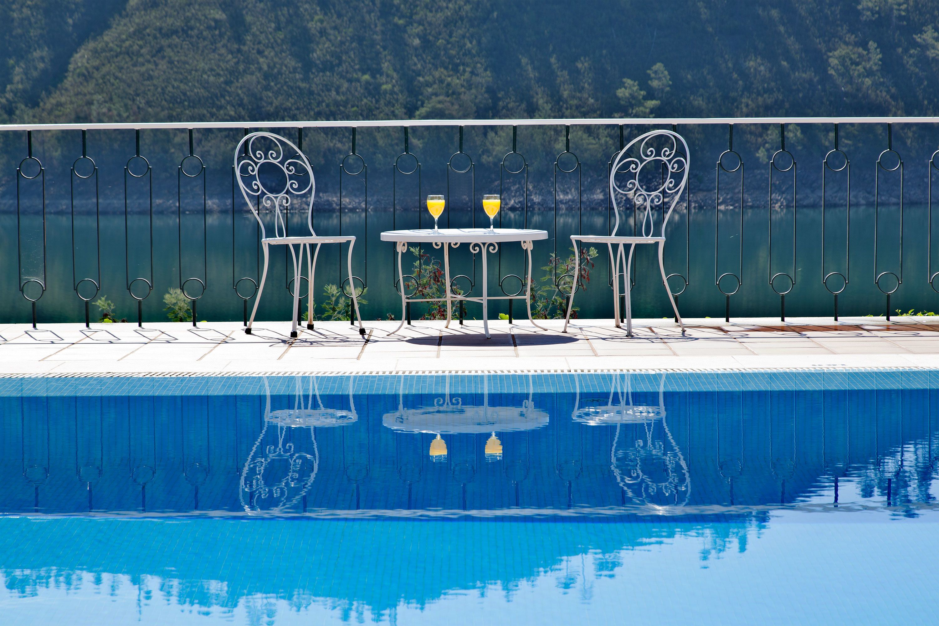 Zona piscina con due sedie, un tavolo, due bicchieri di una bevanda gialla e vista su un pendio boscoso.