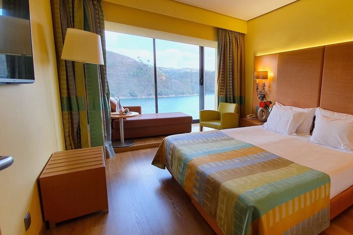 Ett hotellrum med en stor säng, ett skrivbord, en stol och utsikt över en sjö eller ett annat vattendrag.
