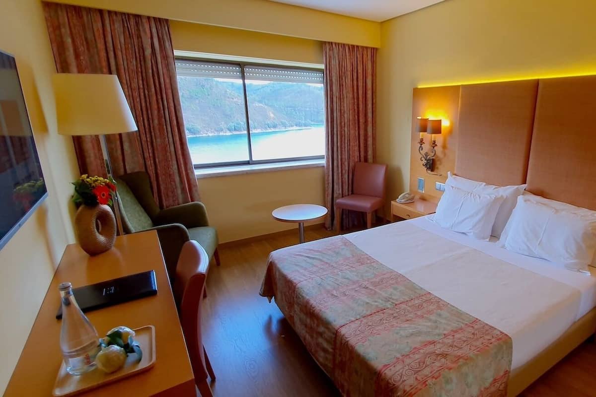Ett hotellrum med en säng, ett skrivbord, en stol, ett litet bord och ett fönster med gardiner.