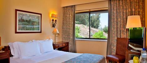 A hotel room with a bed, a desk with a computer, a chair, a window with a view of greenery, and a painting on the wall.