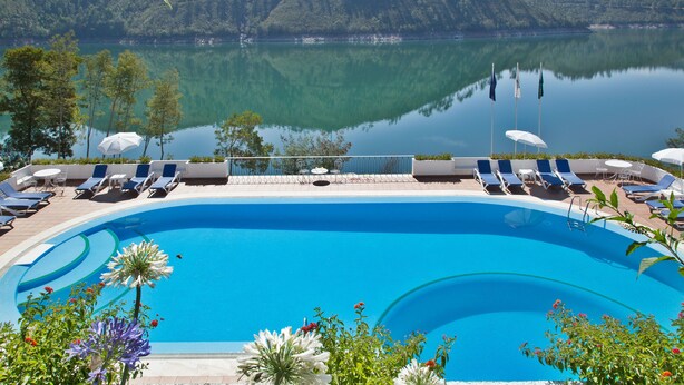 Una piscina con sillones de descanso y sombrillas, rodeada de vegetación y un lago con montañas al fondo.