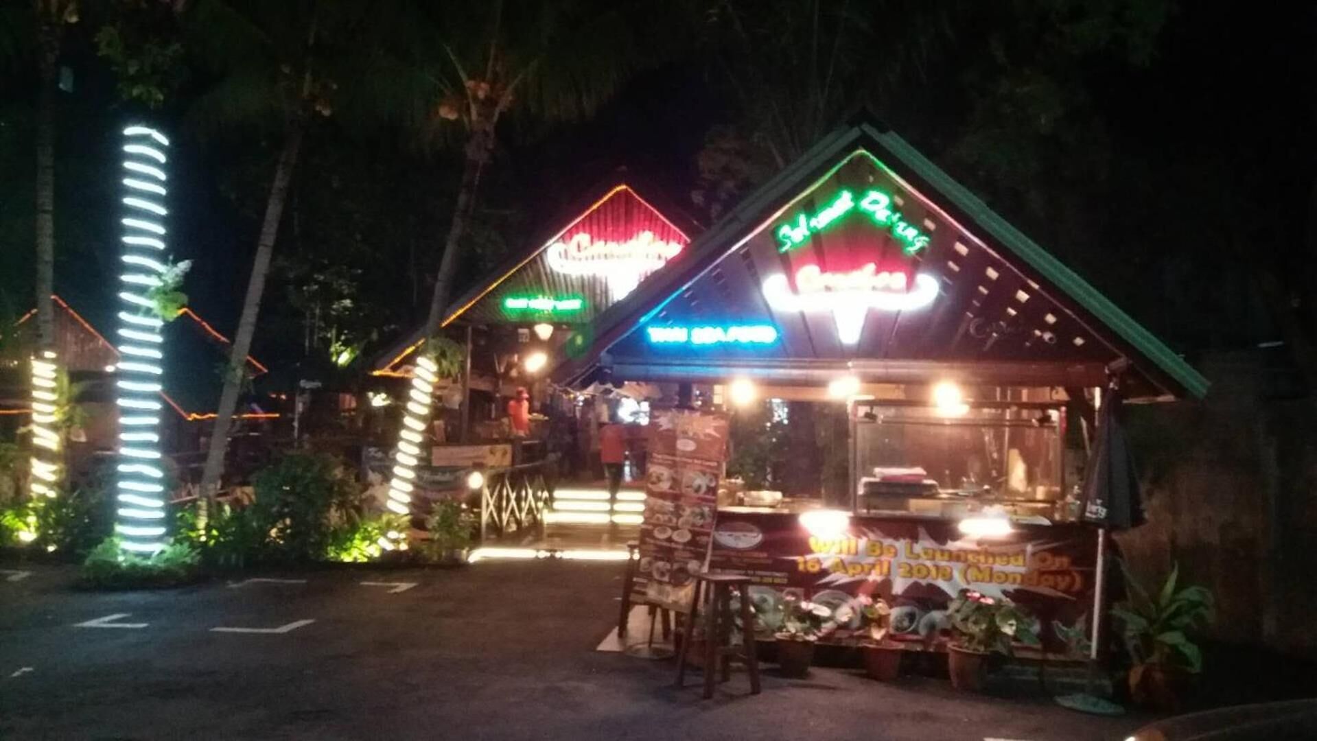 A small, brightly lit outdoor food stall with a neon sign, surrounded by trees and a tall, illuminated structure.