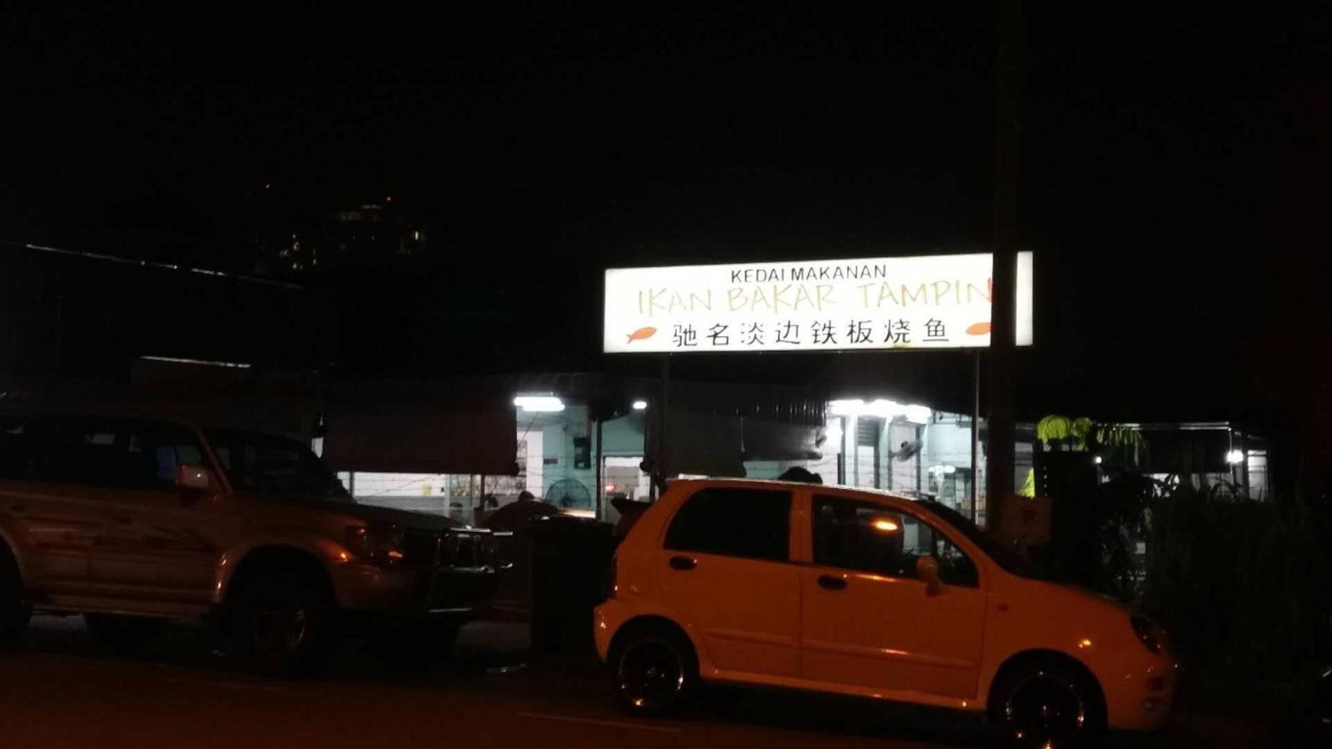 A nighttime street view with a restaurant named 'Ikan Bakar Tampin' and a white car parked in front.