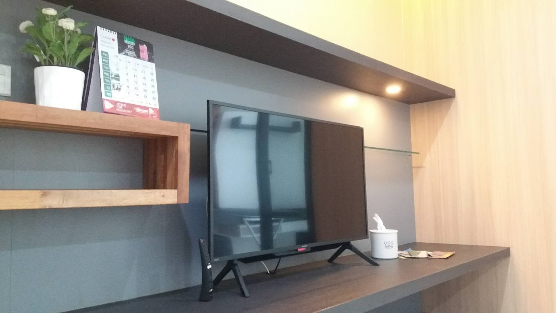 A flat-screen TV mounted on a wall with a wooden shelf above it, a potted plant, and a calendar.