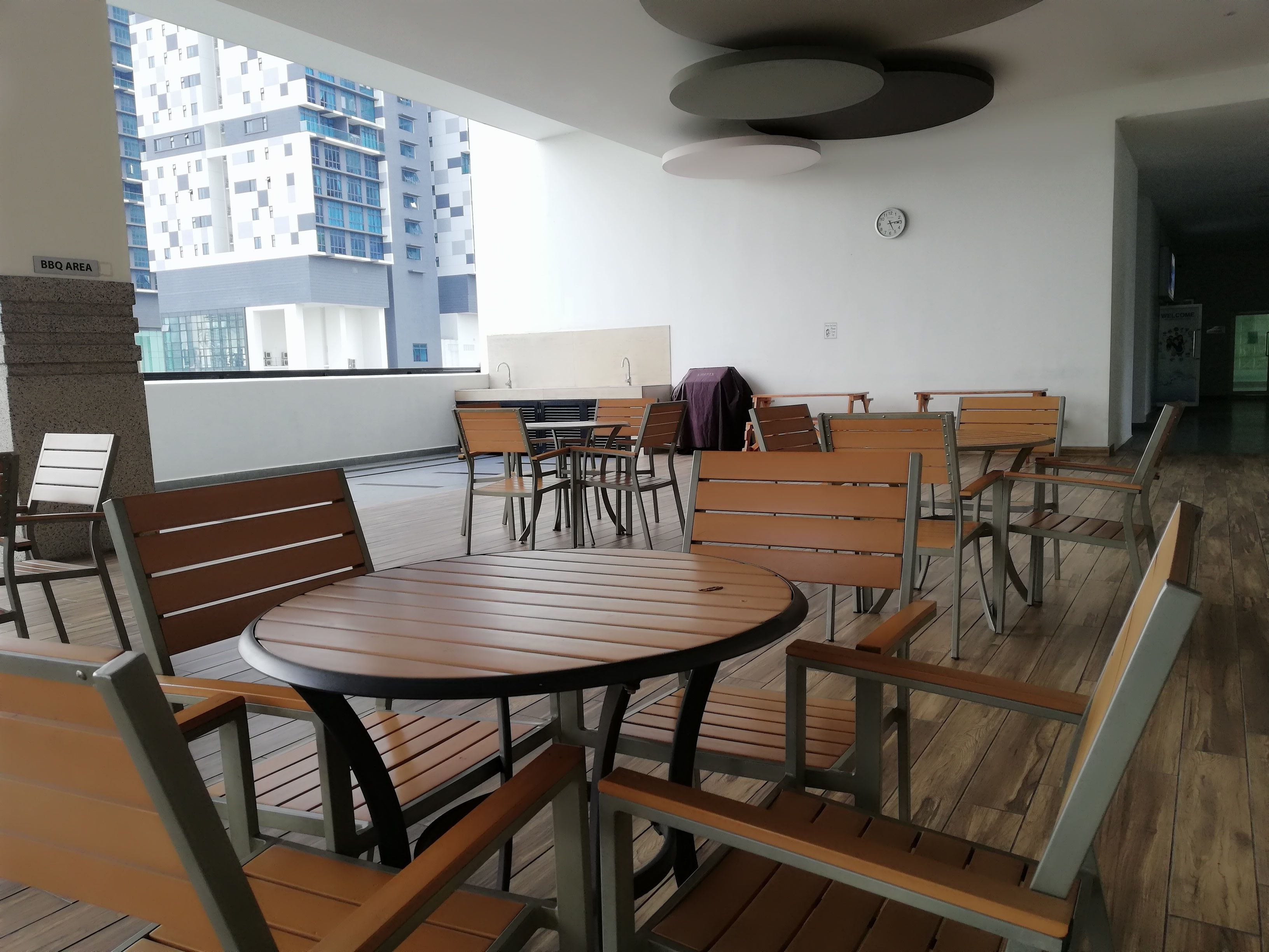 A modern indoor seating area with round tables and wooden chairs, a clock on the wall, and a view of a high-rise building.