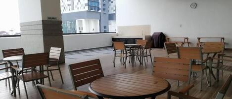 A rooftop terrace with wooden tables and chairs, a view of a modern building, and a clock on the wall.