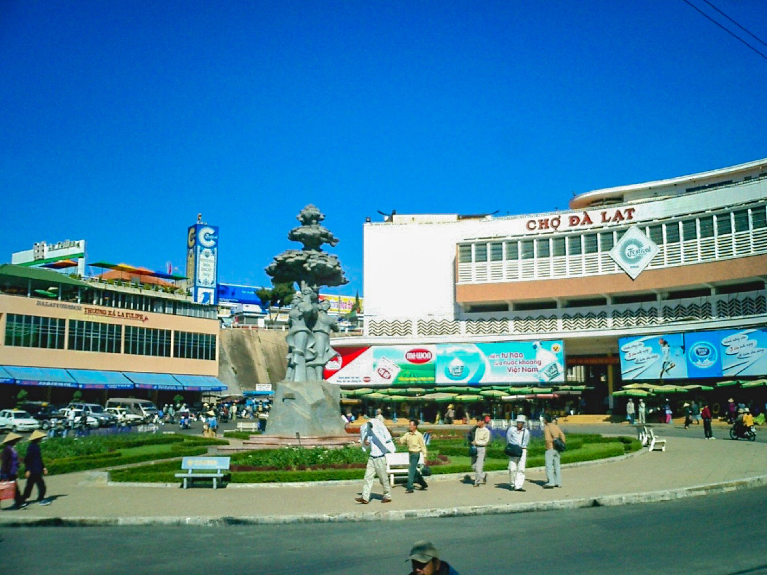 A public square with a central fountain, surrounded by a well-maintained garden and a busy street with pedestrians and vehicles.