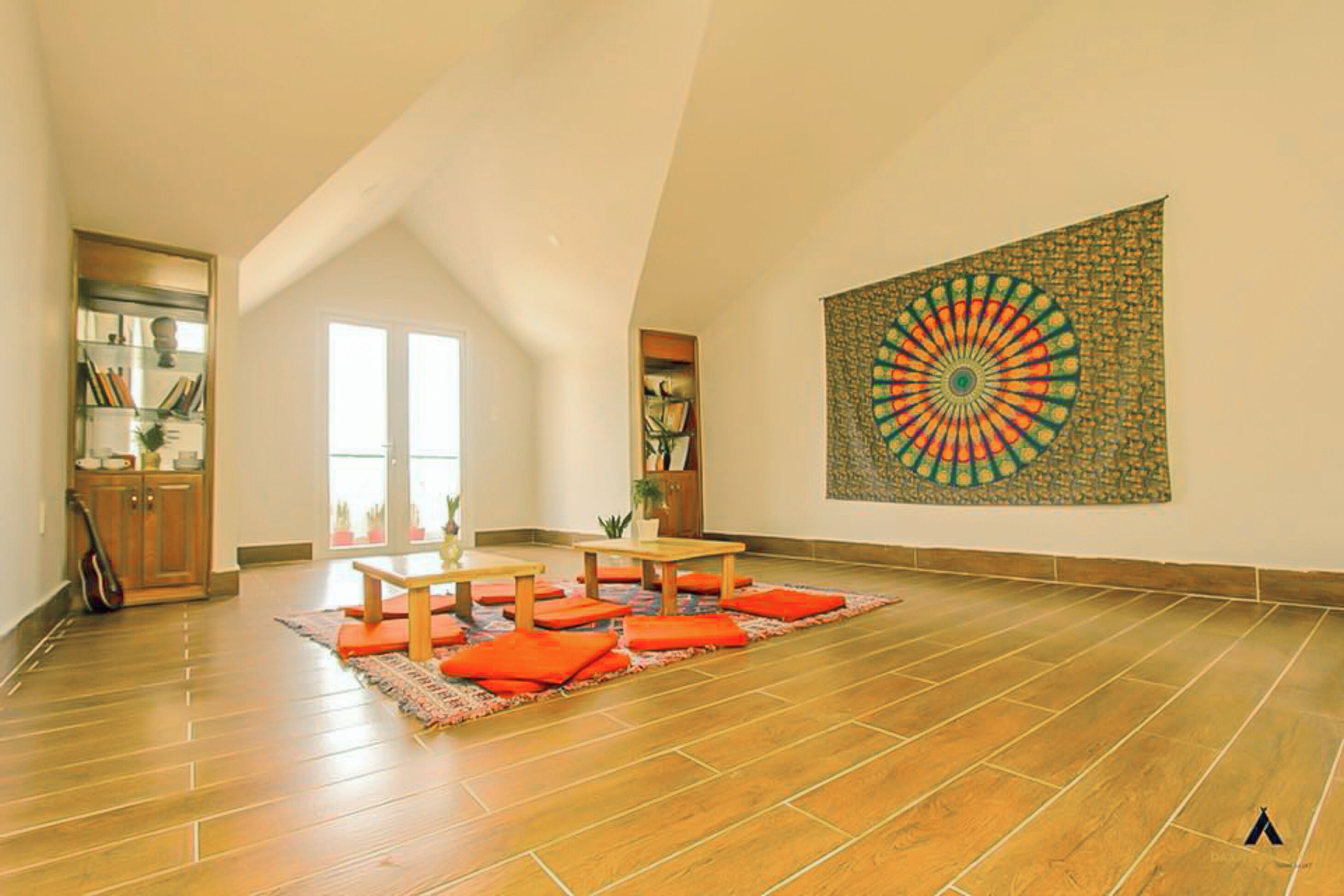 Una habitación amplia con techo alto, un ventanal grande, piso de madera, una mesa de comedor con cojines naranjas y un tapiz colorido en la pared.