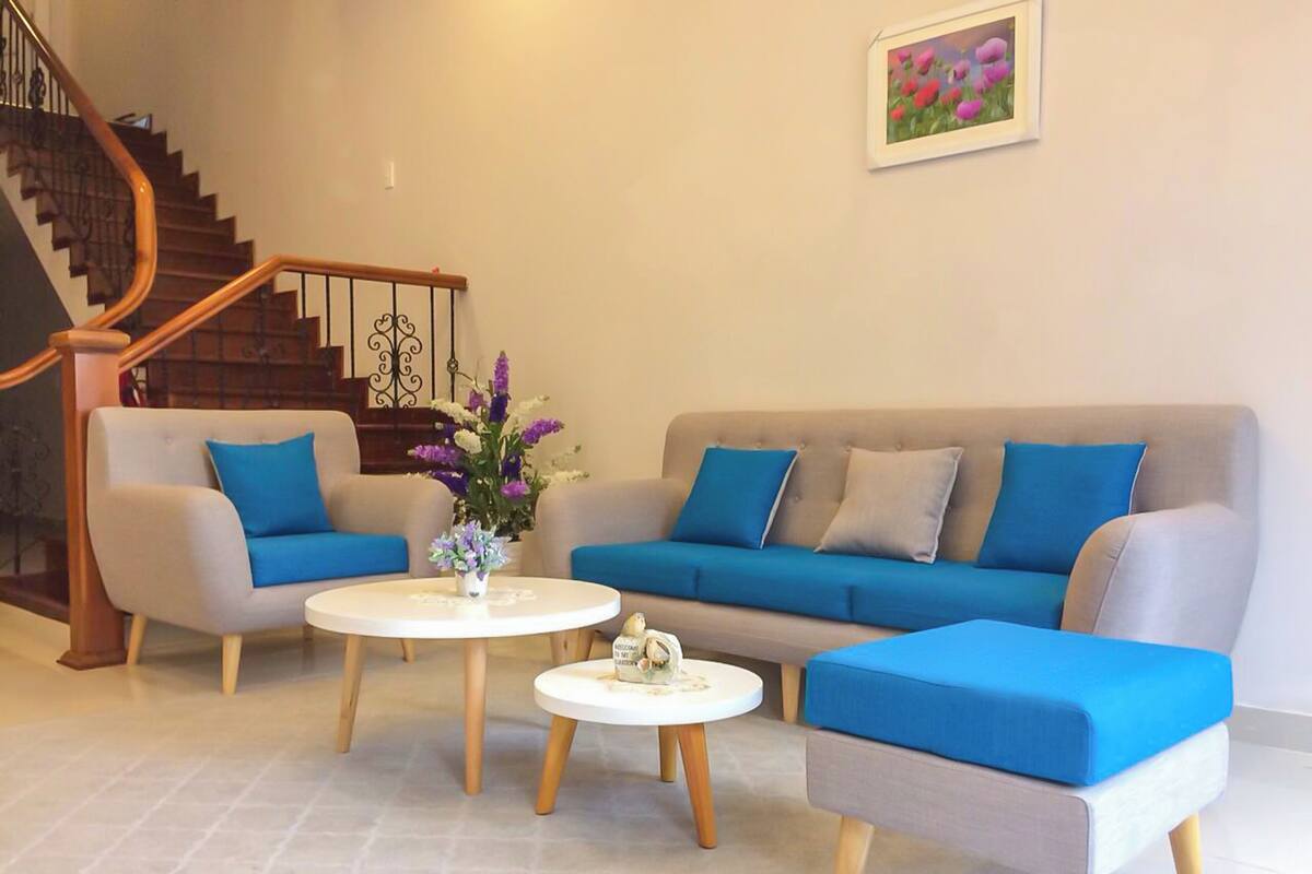 A living room with a grey sofa, two armchairs, a coffee table, and a small table with a vase of flowers. A staircase is visible in the background.
