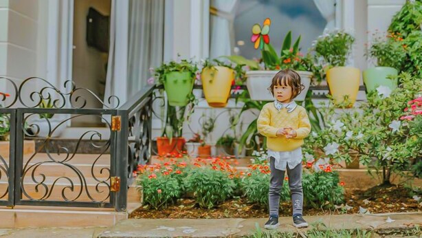 A child standing in front of a building entrance with a metal gate, surrounded by potted plants and flowers. - Jolie House (Lam Vien - Da Lat)