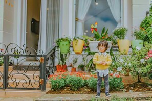 A child standing in front of a building entrance with a metal gate, surrounded by potted plants and flowers. - Jolie House (Lam Vien - Da Lat)