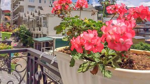 A balcony with a potted plant of pink flowers, a metal railing, and a building with multiple windows in the background. - Jolie House (Lam Vien - Da Lat)