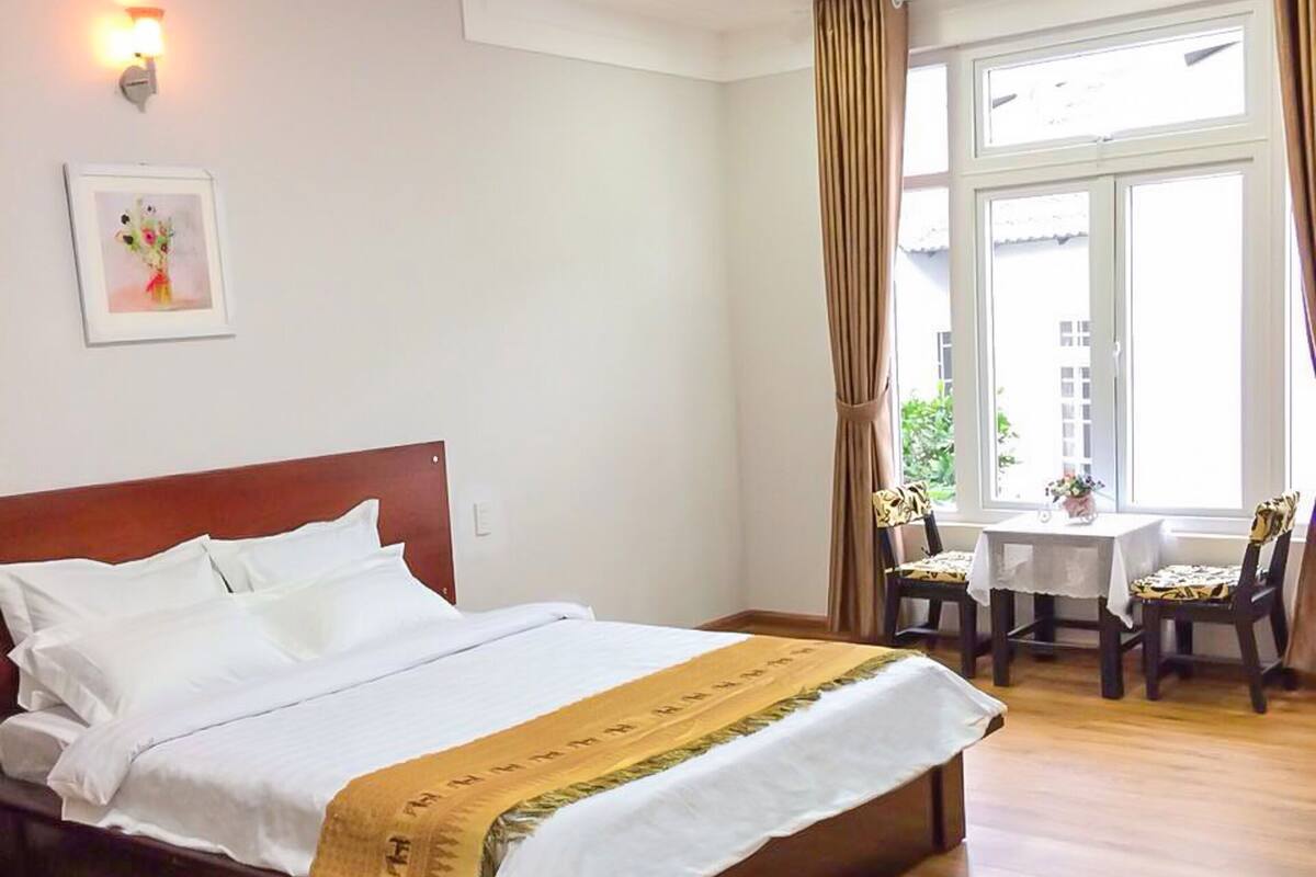 A neatly made bed with a white comforter and a brown blanket, a small table with a white tablecloth, and a chair with a patterned cushion.