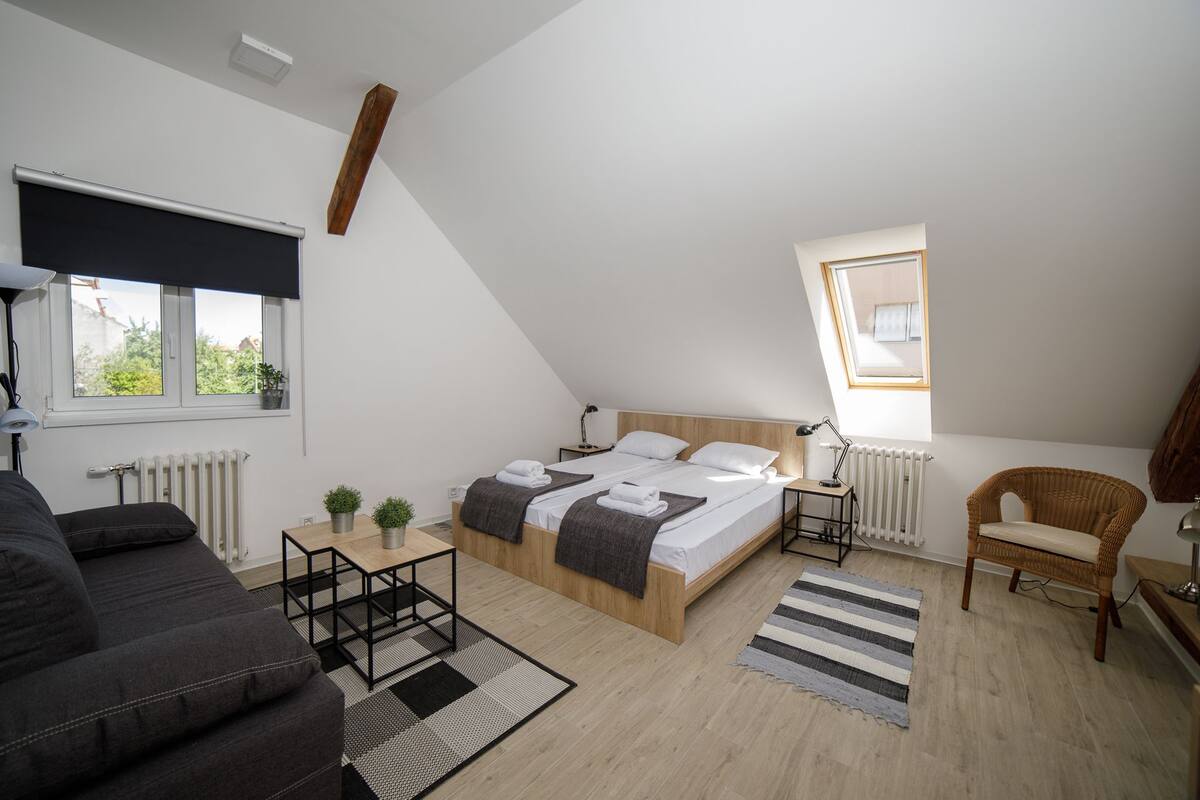 A modern, well-lit bedroom with a large bed, a small table, a chair, and a view of greenery outside.