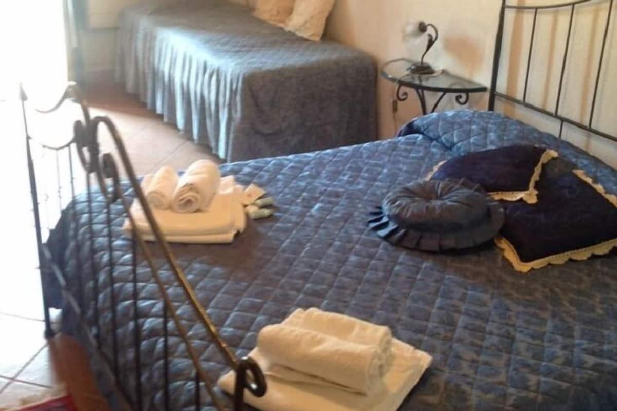 A room with two beds, one with a blue quilt and the other with a grey one. There are towels on the beds and a small table with a lamp.