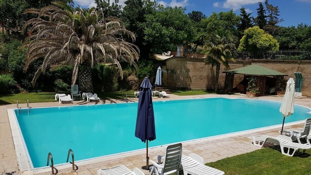 Una piscina rectangular d'aigua clara i blava, envoltada per un paviment de pedra. Hi ha diverses gandules blanques i ombrel·les al voltant de la piscina.