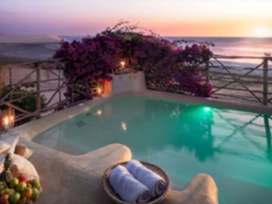 Zona junto a la piscina con vista al océano al atardecer, rodeada de una terraza de madera y flores moradas.