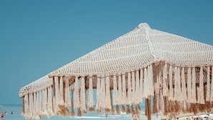 A beach scene with a couple relaxing under a macrame umbrella, a lounge chair, and a table with drinks. - Casa de Areia (Jijoca de Jericoacoara)