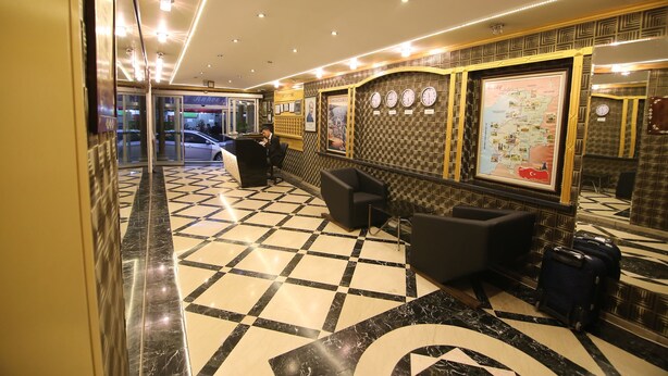 A hotel lobby with a checkered floor, a reception desk, a seating area, and a wall with a map and clocks. - Aykut Palace Otel (Iskenderun)