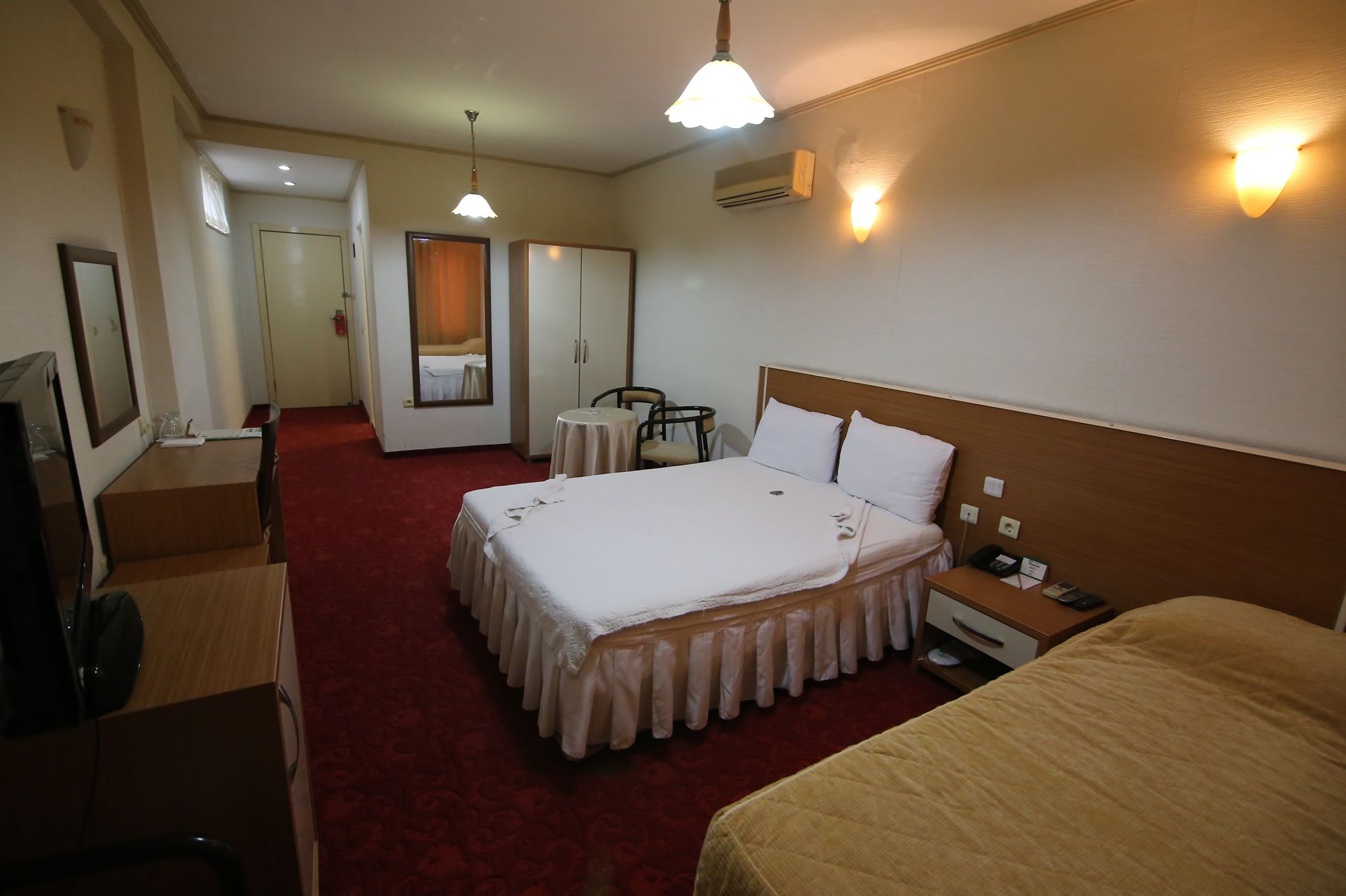 A hotel room with a large bed, a smaller bed, a desk with a chair, a mirror, and a television.