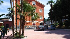 A multi-story building with a red facade, palm trees, and a paved area with parked cars. - Hotel Locanda l'Ombrosa (Vezzano Ligure)